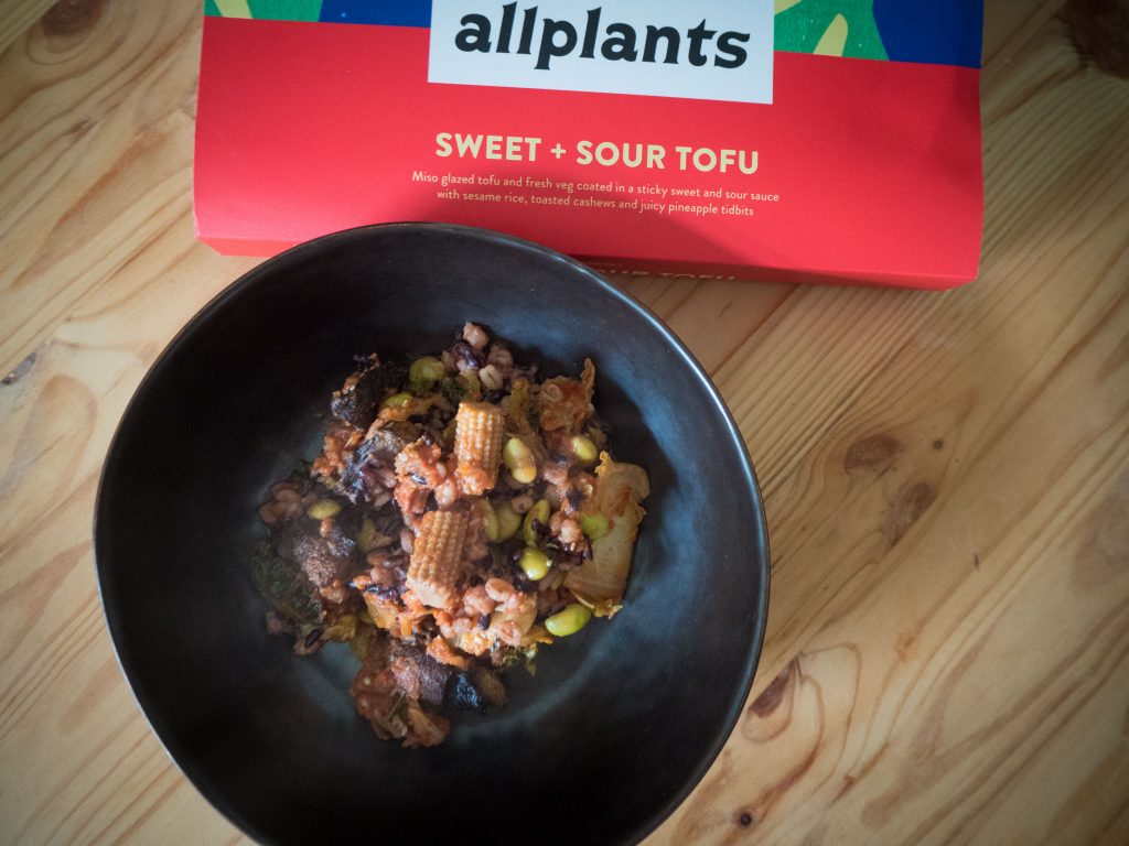 allplants sweet and sour tofu plated in a black bowl next to it's red packaging